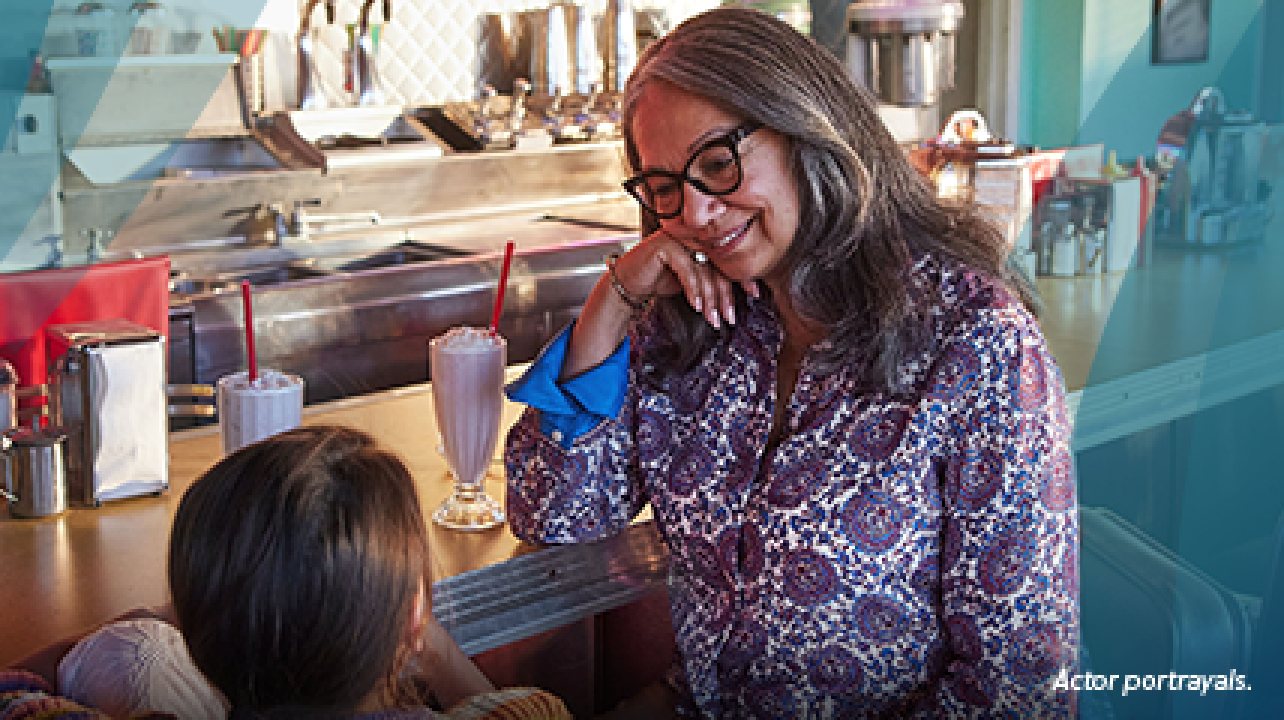 A Latina woman in her 60s and her young granddaughter are sitting on barstools at a vintage diner counter, looking at each other and smiling. The woman has long gray hair and bold glasses and is resting her elbow on the counter. Two strawberry milkshakes with whipped cream and long red straws are on the counter beside them. Actor portrayals.