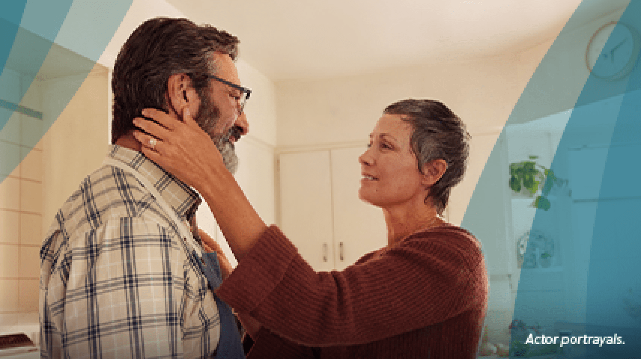 A white woman in her 50s with short gray hair is standing in the kitchen with her partner, a white man in his 50s. The woman is reaching out, with one hand resting on the man's shoulder and the other hand gently holding the man's jaw. They are looking at each other fondly. Actor portrayals.