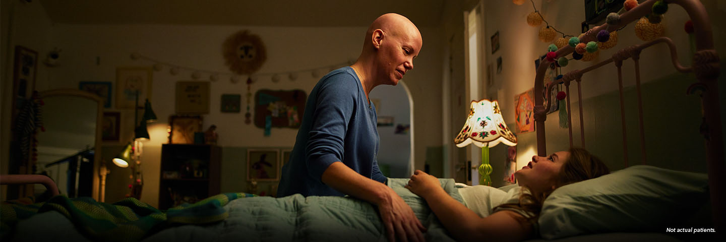 A white woman in her early 40s with no hair is sitting on the side of her young daughter's bed, tucking her in for the night. They are softly smiling at each other. Not actual patients.