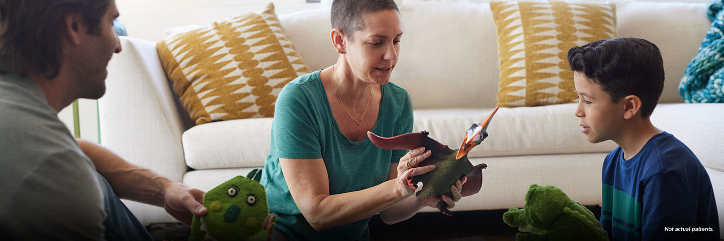 A white woman in her 30s with shaved dark hair sits on the living room floor in front of the couch between a white man in his 30s and a 7-year-old boy. The woman is holding a dinosaur action figure and is playing with the boy, who has a dinosaur puppet on his hand. Not actual patients.