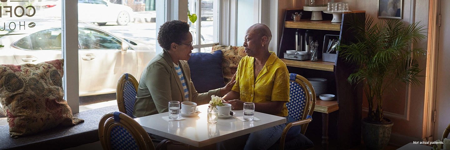 A Black woman in her 40s with closely shaved hair sits at a table in a coffee shop with her friend, another Black woman in her 40s, who is gently holding the other woman's arm in a gesture of support. Not actual patients.
