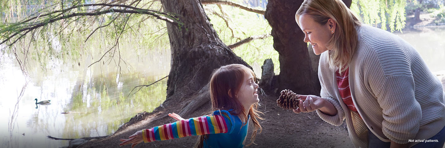 A white woman in her mid-30s with shoulder-length, blonde, straight hair is out on a nature walk with her 6-year-old daughter. The woman is smiling and holding a pinecone that her daughter found, as the girl smiles and jumps excitedly. Not actual patients.