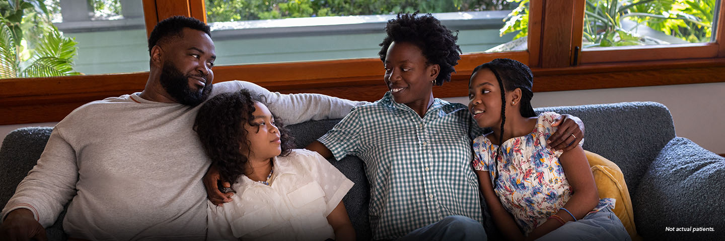 A Black woman in her mid-40s with short natural hair is cuddled on the living room couch with her family--a Black man in his 40s and their 2 preteen daughters. The woman has her arms around both of her daughters and is smiling at her husband. The family is close, leaning on each other and smiling. Not actual patients.