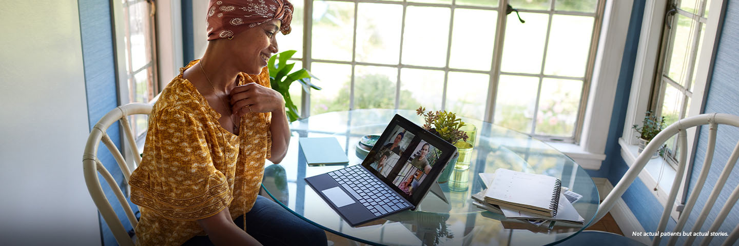 A Black woman in her early 40s wearing a paisley headscarf and a patterned yellow shirt sits in a brightly lit room at a round glass table smiling at her tablet screen, on a video call with her close group of friends. A potted plant, notebook, and notepad sit on the table with her tablet. Not actual patients, but actual stories.