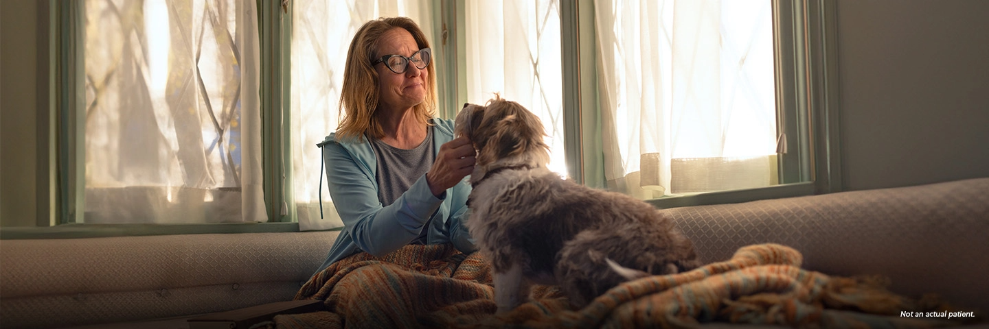 A white woman in her 50s with shoulder-length blond hair and glasses is sitting on her couch and smiling softly as she pets a medium-sized tan and white dog with shaggy fur. Not an actual patient.
