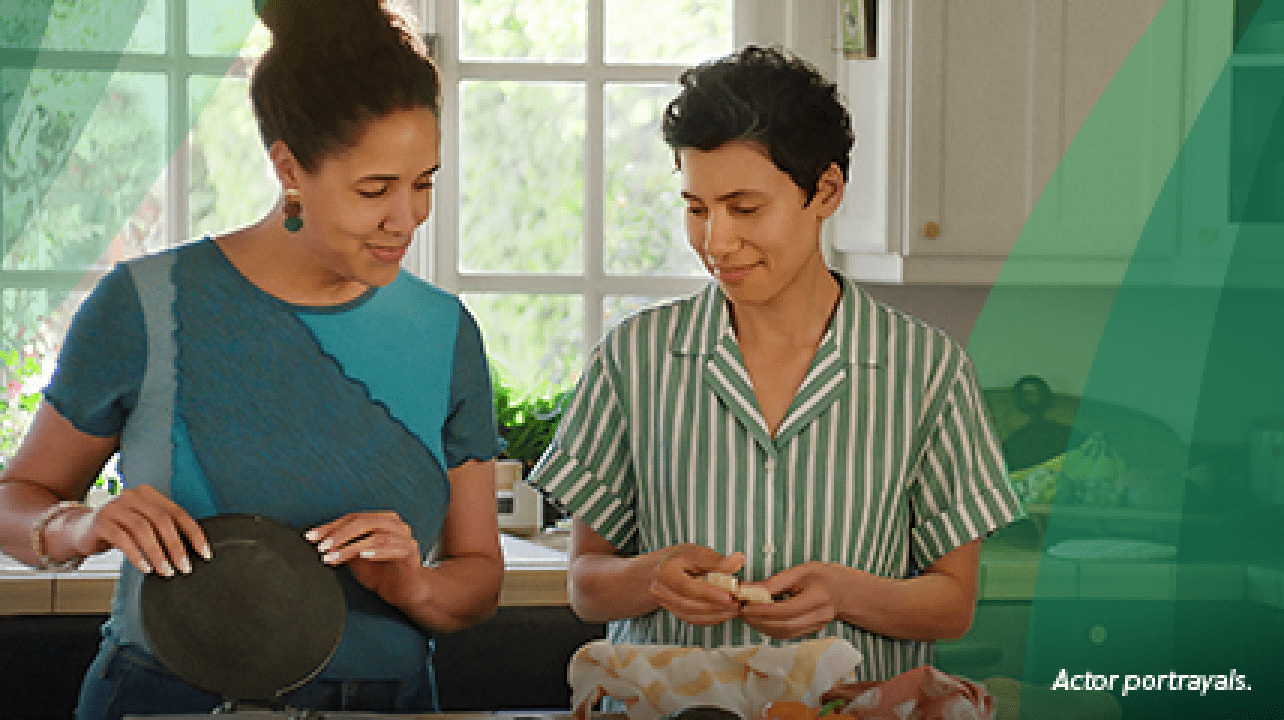 A Latina woman in her mid-40s with short, dark, curly hair stands in the kitchen with her partner, another Latina woman in her 40s. They are making tortillas from scratch, with one woman forming the masa and the other pressing it in the tortilla press. Actor portrayals.