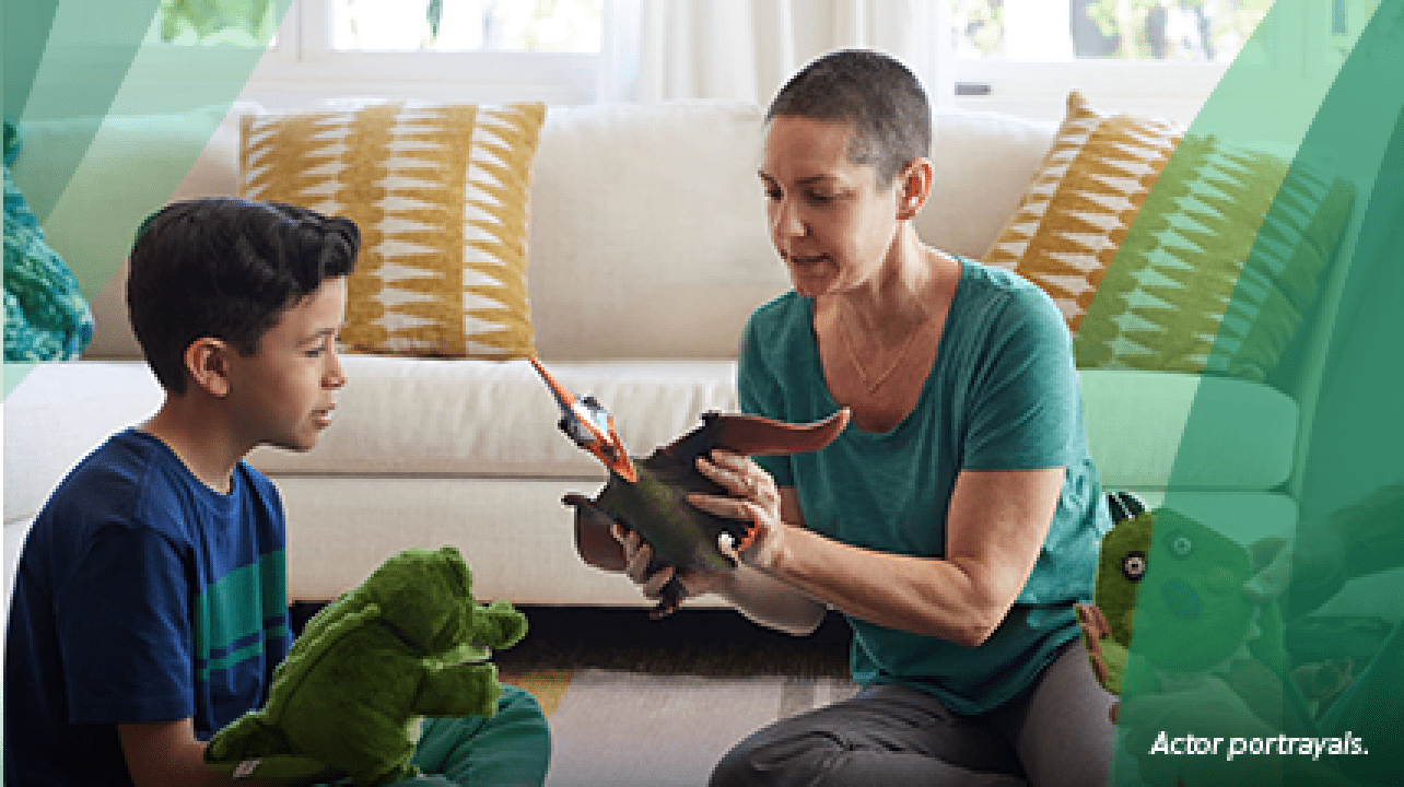 A white woman in her 30s with shaved dark hair sits on the living room floor in front of the couch between a white man in his 30s and a 7-year-old boy. The woman is holding a dinosaur action figure and is playing with the boy, who has a dinosaur puppet on his hand. Actor portrayals.