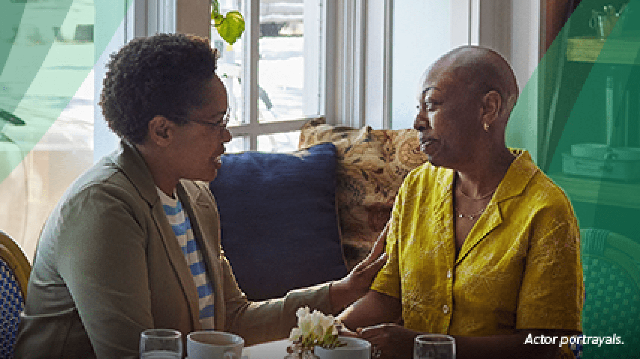 A Black woman in her 40s with closely shaved hair sits at a table in a coffee shop with her friend, another Black woman in her 40s, who is gently holding the other woman's arm in a gesture of support. Actor portrayals.