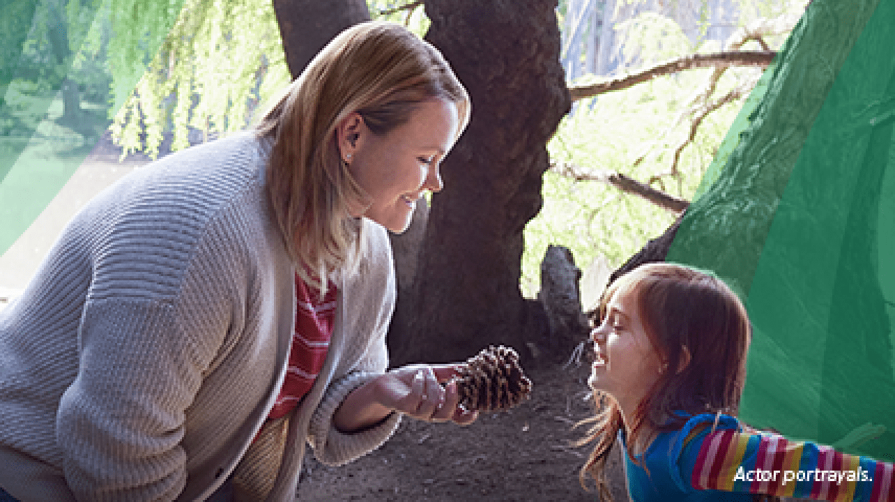 A white woman in her mid-30s with shoulder-length, blonde, straight hair is out on a nature walk with her 6-year-old daughter. The woman is smiling and holding a pinecone that her daughter found, as the girl smiles and jumps excitedly. Actor portrayals.