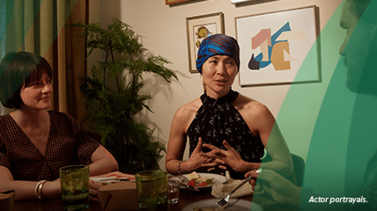 An Asian woman in her early 40s wearing a red and blue silk headscarf sits at a round dining table, smiling and talking with 2 friends, a white man and white woman of a similar age. They are finishing dinner, a takeout meal from one of their favorite restaurants. Actor portrayals.