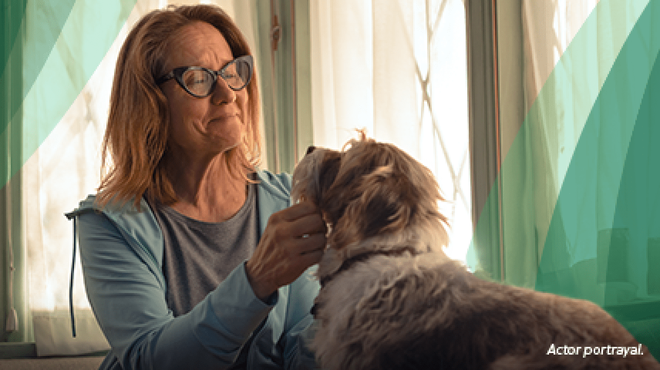 A white woman in her 50s with shoulder-length blond hair and glasses is sitting on her couch and smiling softly as she pets a medium-sized tan and white dog with shaggy fur. Actor portrayal.