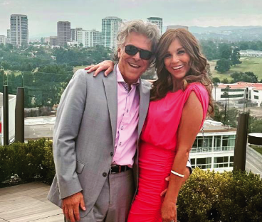Jacquie, a woman receiving TRODELVY, is standing with her arm around her husband, Marc. Jacquie is a White woman, wearing a red dress and a wig. Marc is a White man with gray hair, wearing a gray suit and sunglasses.