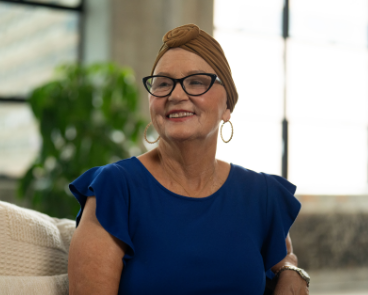 Jana, a woman receiving TRODELVY, sits smiling on a couch. Jana is a White woman wearing a headscarf, glasses, hoop earrings, and a blue shirt.
