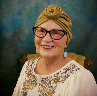 Jana, a woman receiving TRODELVY, sits smiling at the camera. Jana is a White woman wearing a headscarf, glasses, and a white patterned shirt.