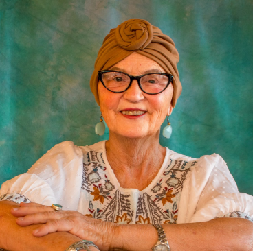Jana, a woman receiving TRODELVY, sits smiling at the camera. Jana is a White woman wearing a headscarf, glasses, and a white patterned shirt.
