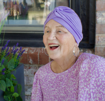 Jana, a woman receiving TRODELVY, sits smiling at the camera. Jana is a White woman wearing a purple headscarf, gold earrings, and a purple floral‐patterned shirt.