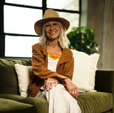 Karen, a woman receiving TRODELVY, is smiling at the camera. Karen is a White woman wearing a blonde wig, a brown sun hat, a brown leather jacket, and a white shirt and white pants.