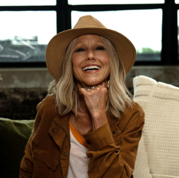 Karen, a woman receiving TRODELVY, is smiling at the camera. Karen is a White woman wearing a blonde wig, a brown sun hat, a brown leather jacket, and a white shirt and white pants.