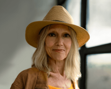 Karen, a woman receiving TRODELVY. Karen is a White woman wearing a blonde wig, a brown sun hat, a brown leather jacket, and a white shirt.