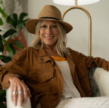Karen, a woman receiving TRODELVY, is smiling at the camera. Karen is a White woman wearing a blonde wig, a brown sun hat, a brown leather jacket, and a white shirt and white pants.