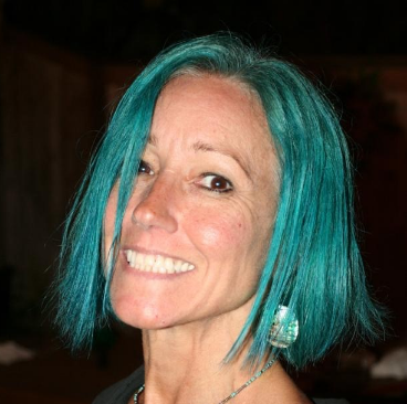 Karen, a woman receiving TRODELVY, is standing outside smiling at the camera. Karen is a White woman wearing a blue wig, and a black shirt.