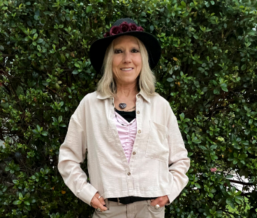 Karen, a woman receiving TRODELVY, is smiling at the camera. Karen is a White woman wearing a blonde wig, a black sun hat with roses on the brim, a tan jacket, a pink shirt, and brown pants.
