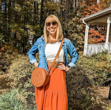 Katherine, a woman receiving TRODELVY, is standing outside smiling at the camera. Karen is a White woman wearing a blonde wig, sunglasses, a denim jacket, a white shirt, an orange skirt, a leather purse, and brown leather boots.