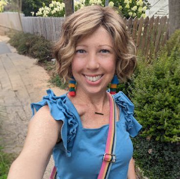 Katherine, a woman receiving TRODELVY, is standing outside and smiling at the camera. Karen is a White woman wearing a blonde wig and a blue shirt.