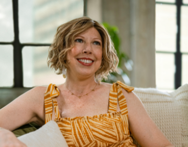 Katherine, a woman receiving TRODELVY, sits smiling at the camera. Katherine is a White woman wearing a blonde wig and a striped dress.