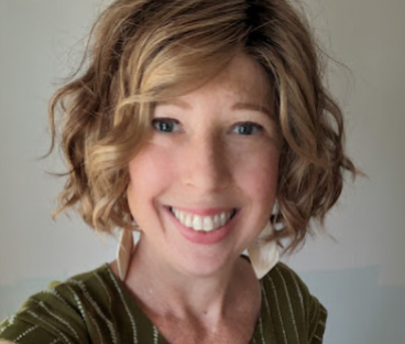Katherine, a woman receiving TRODELVY, is smiling at the camera. Katherine is a White woman wearing a blonde wig, and a brown shirt with vertical white stripes.