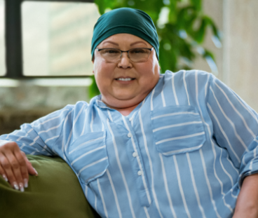 Lisa, a woman receiving TRODELVY, sits smiling at the camera. Lisa is a Latina woman wearing a headscarf, glasses, and a blue shirt with white stripes.