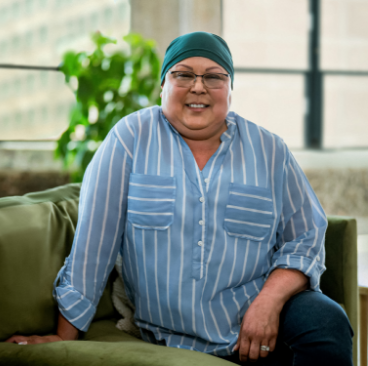 Lisa, a woman receiving TRODELVY, sits smiling at the camera. Lisa is a Latina woman wearing a headscarf, glasses, and a blue shirt with white stripes.