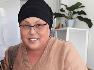 Lisa, a woman receiving TRODELVY, sits smiling at the camera. Lisa is a Latina woman wearing a headscarf, glasses, and a tan shirt.