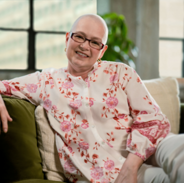 Tanya, a woman receiving TRODELVY, sits smiling at the camera. Tanya is a White woman with no hair, wearing glasses and a white shirt with a red floral pattern.