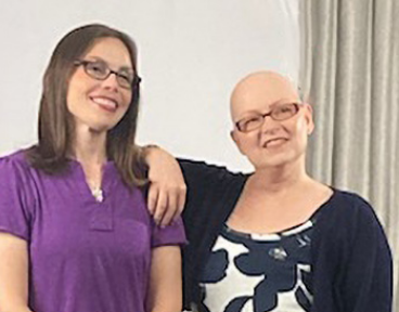 Tanya, a woman receiving TRODELVY, stands with her care partner, both smiling at the camera. Tanya is a White woman with no hair, wearing glasses and a black and white dress with a black jacket. Tanya's care partner is a White woman with brown hair, wearing glasses, a purple shirt, and black pants.