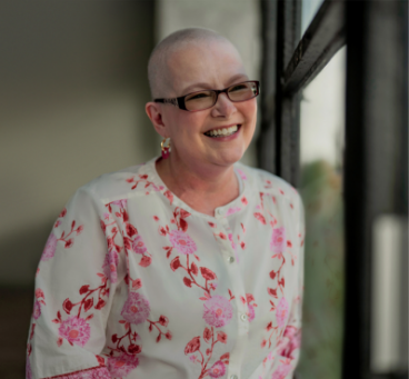 Tanya, a woman receiving TRODELVY, sits smiling at the camera. Tanya is a White woman with no hair, wearing glasses and a white shirt with a red floral pattern.
