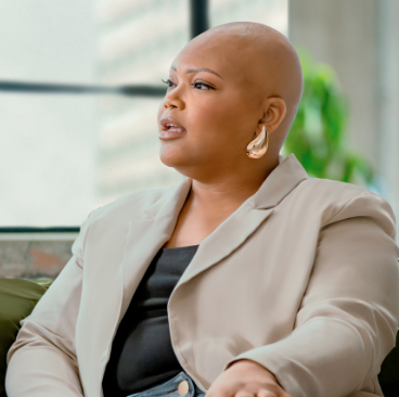 Tiana, a woman receiving TRODELVY, is sitting on a couch. Tiana is a Black woman with no hair, wearing a black shirt under a tan jacket, and denim jeans.