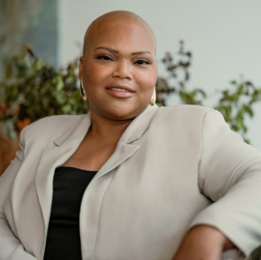 Tiana, a woman receiving TRODELVY, is sitting on a couch while smiling at the camera. Tiana is a Black woman with no hair, wearing gold earrings, and a black shirt under a tan jacket.