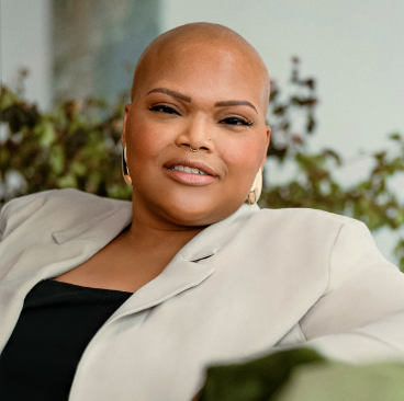 Tiana, a woman receiving TRODELVY, is sitting on a couch while smiling at the camera. Tiana is a Black woman with no hair, wearing gold earrings, and a black shirt under a tan jacket.