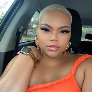 Tiana, a woman receiving TRODELVY, sits in her car smiling at the camera. Tiana is a Black woman with short blonde hair wearing an orange shirt, gold earrings, and a gold watch.