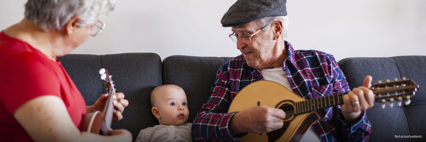 A white man and woman in their 70s are sitting on a couch with a baby. The woman is playing a ukulele and the man is playing a mandolin and they are singing happily to the baby.