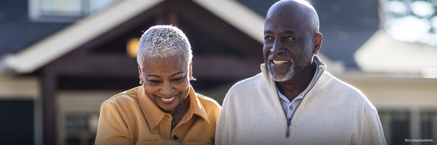 A Black man and woman in their 60s are outside with a house in the background. They are walking together and smiling softly.