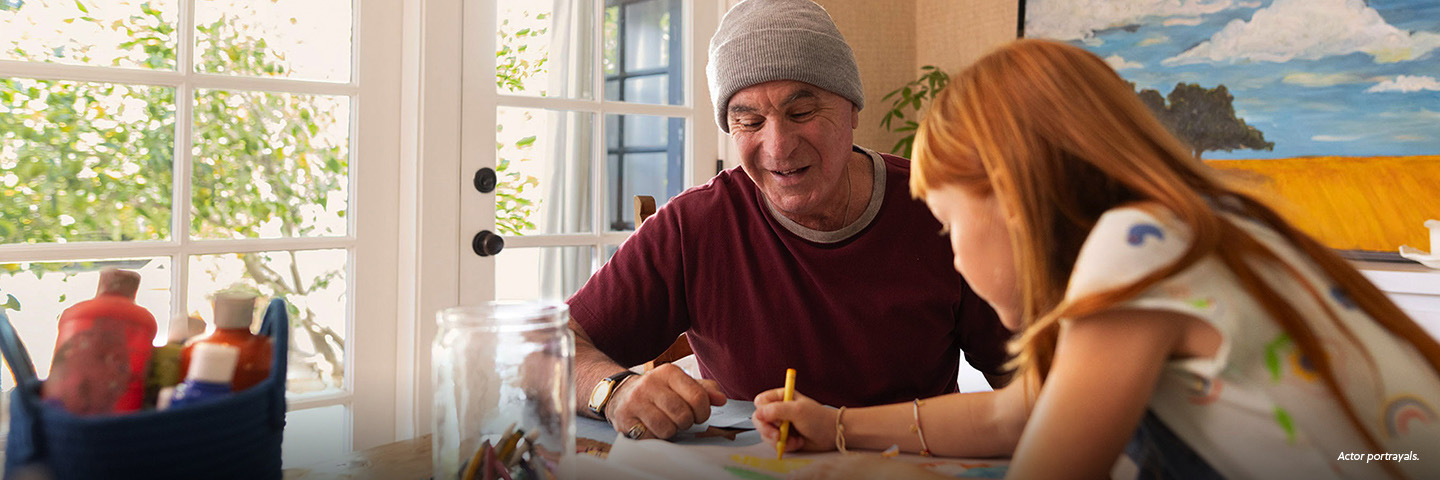 A White man in his 60s is sitting at a table with his redheaded granddaughter. They are coloring on paper with different colored crayons, softly smiling. Actor portrayals.
