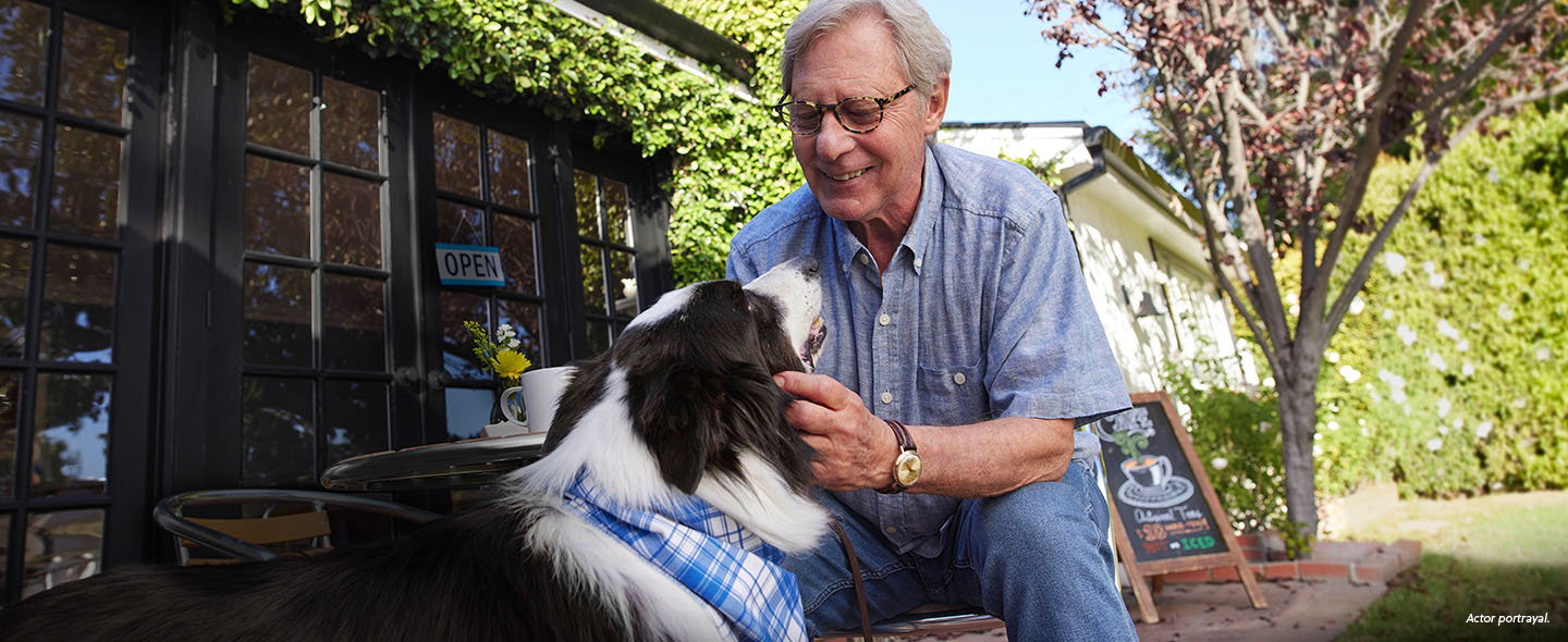 A White man in his 70s is outside of a coffee shop, kneeling down and petting a black and white dog that is looking up at the man. The man is smiling at the dog. Actor portrayal.