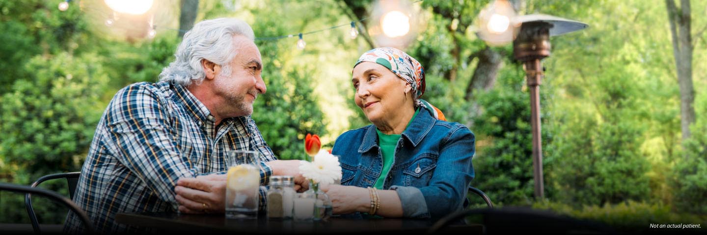 A Hispanic man and woman in their 60s are sitting at a dining table on an outdoor patio. She is wearing a floral head scarf and they are looking at each other and smiling softly. Not actual patients, but actual stories.